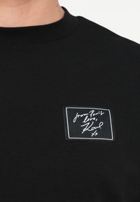 Černý crewneck top s malou obdélníkovou nášivkou na hrudi, na které je bíle psáno "from Paris love, Karl xx" v kurzívě.