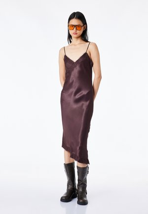 Nainen, jolla on päällään tummanvioletti satiininen slip-mekko, jossa on pitsireunus, oranssisävyiset aurinkolasit ja mustat puolivälin varren saappaat, seisoo valkoisen taustan edessä.