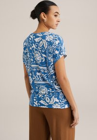 Zils ziedu raksts tops ar īsām piedurknēm un brīvu piegriezumu, ar baltām botāniskām dizainu detaļām un gludu tekstūru.