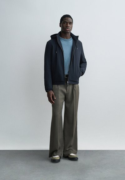 Άνδρας που φοράει μαύρο μπουφάν με κουκούλα πάνω από μπλε πουλόβερ, φαρδιά καφέ παντελόνια και μπεζ-μαύρα παπούτσια, στέκεται σε απλό γκρι φόντο.