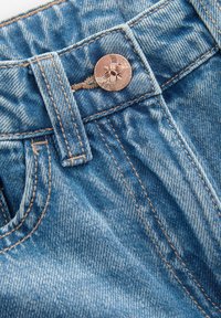Gros plan sur un jean en denim bleu montrant un bouton en laiton, une boucle de ceinture et des coutures beiges détaillées sur la ceinture et la poche.