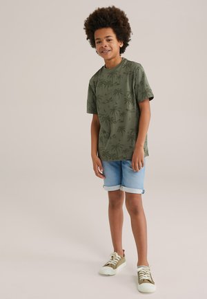 T-shirt verde a maniche corte con stampa di palme, abbinato a pantaloni corti in denim chiaro e sneakers beige. Tessuto morbido, vestibilità rilassata.