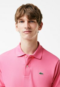 Rosa polo shirt i texturerat tyg, med klassisk krage, två knappar och en grön krokodil-logotyp på vänster bröst.