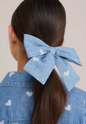 Păr șaten închis prins în spate cu o fundă mare albastră cu modele albe în formă de inimă, asortată cu cămașa albastră cu inimioare albe.