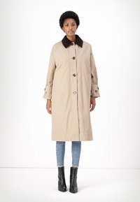 Beige színű trench coat barna gallérral, elöl gombos záródással és oldalsó zsebekkel; kék farmernadrág és fekete bokacsizma felett viselve.