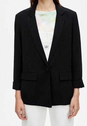 Person iført en sort enkeltknappet blazer over en lys batikfarvet skjorte og hvide bukser, stående med hænderne afslappet langs siderne.