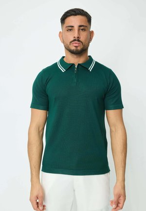Homme aux cheveux courts et foncés et à la barbe, portant un polo à manches courtes zippé de couleur vert foncé et un pantalon blanc, se tenant devant un fond uni.