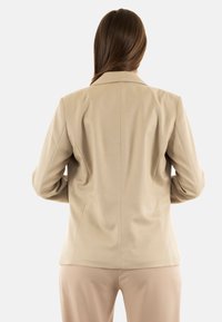 Blazer beige a medida con cuello de muesca, textura suave y ajuste estructurado, con detalles de costura y mangas largas.