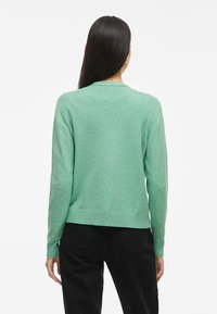 Suéter verde de malha com decote redondo, bainha e punhos canelados, apresentando uma cor sólida e textura suave, usado sobre calças pretas.