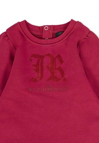 Felpa fucsia con maniche a sbuffo, caratterizzata da un logo "R" testurizzato e la scritta "JOHN RICHMOND" stampata sotto. Chiusura a snap sulla parte posteriore del collo.