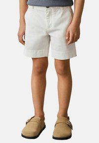 Pantalones cortos de algodón blancos con un frente plano y bolsillos laterales, combinados con zapatos slip-on de ante beige que cuentan con una cinta y suelas negras.