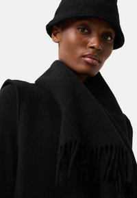 Sort uldtørklæde med frynser på kanterne, lagdelt over en sort frakke. Modellen bærer en sort bucket hat, der fremhæver en glat tekstur og et sømløst design.