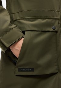 La main d'une personne dans la grande poche à rabat d'une veste vert olive avec une lanière boutonnée au niveau du poignet et un petit patch logo.