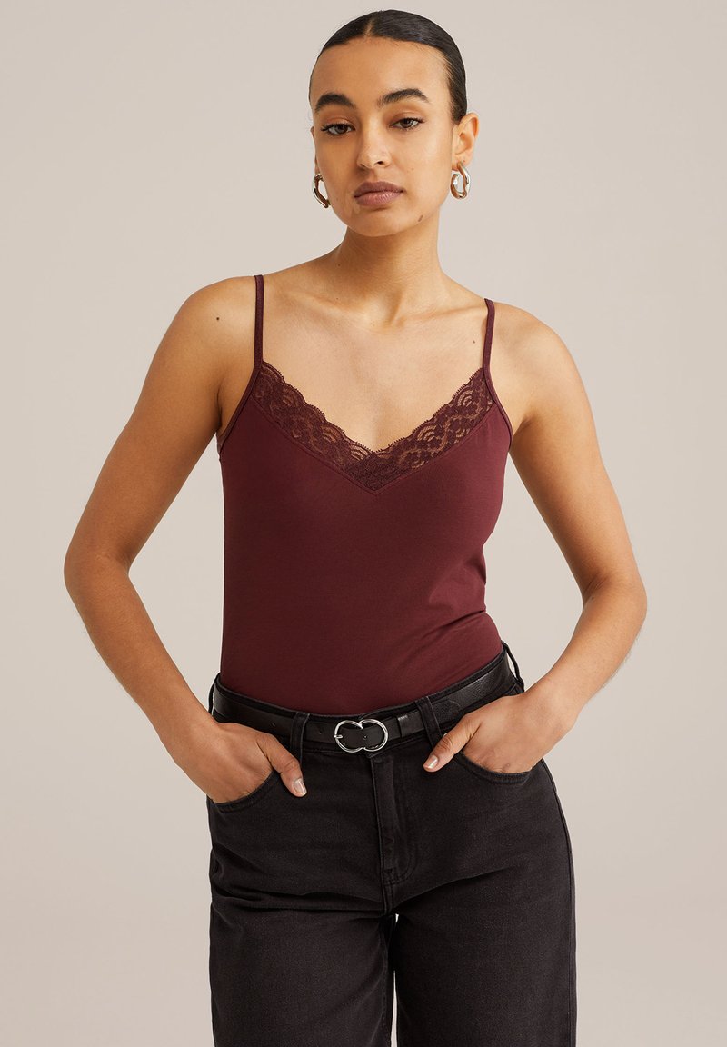 Burgundy tank top dengan hiasan renda, tali spaghetti yang dapat disesuaikan, dan potongan slim, dipadukan dengan celana denim hitam berpinggang tinggi dan sabuk.