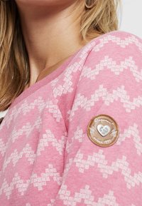 Felpa rosa con un motivo geometrico bianco, scollatura a girocollo e dettaglio distintivo tondo e strutturato sulla manica. Tessuto morbido in misto cotone.