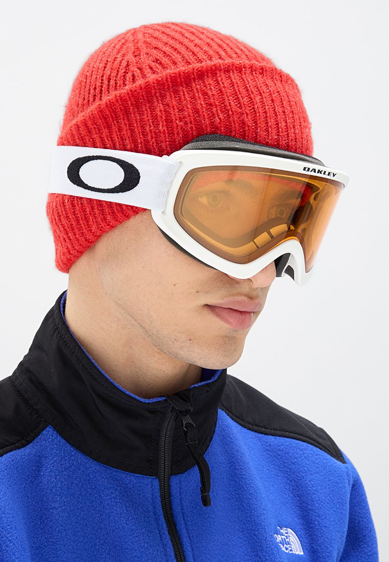 Gafas de esquí Oakley con lentes naranjas y marco blanco, combinadas con un gorro de lana rojo de canalé. El portador lleva una chaqueta de forro polar azul con cuello.