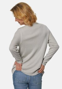 Lichtgrijze ribgebreide top met lange mouwen en een ronde onderrand, voorzien van zijsplitsingen, gecombineerd met blauwe jeans. De achterkant toont details van de textuur van de stof.