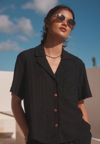 Short-sleeved black shirt with a textured pattern, button closure, and a chest pocket; paired with round sunglasses.