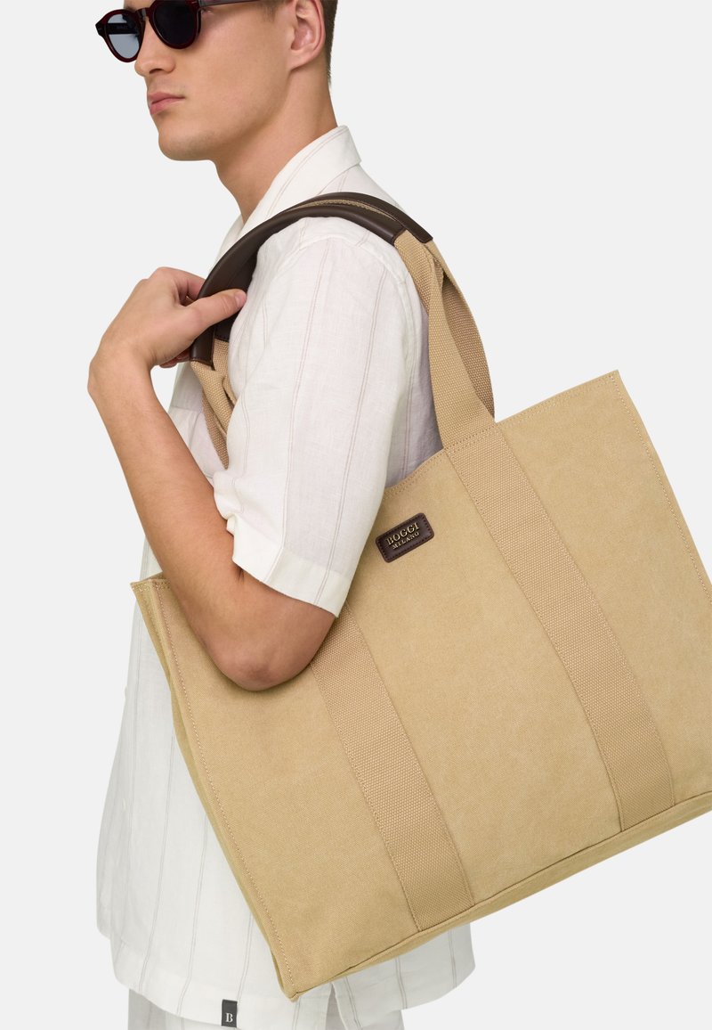 Homme portant une chemise blanche à manches courtes et des lunettes de soleil noires transporte un grand sac fourre-tout en toile beige à poignées en cuir sur l'épaule.