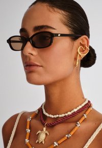 Γυναίκα με μαλλιά χτενισμένα προς τα πίσω φοράει μαύρα ορθογώνια γυαλιά ηλίου, μεγάλο χρυσό σκουλαρίκι-μανσέτα στο αυτί κ αι πολλαπλά κολιέ, μεταξύ των οποίων ένα χρυσό με μενταγιόν σε σχήμα χελώνας.