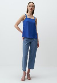 Koninklijk blauwe mouwloze top met brede banden, gecombineerd met lichtblauwe recht gesneden jeans en zilveren sandalen met banden. Glad stoftextuur.