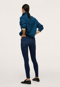 Suéter de punto color teal con un cuello alto, combinado con jeans skinny azul oscuro y mocasines negros. El conjunto presenta un patrón de punto texturizado.