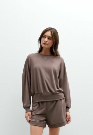 Vrouw met lang bruin haar draagt bijpassende losse bruine sweatshirt en korte broek, staat tegen een effen lichte achtergrond en kijkt opzij.