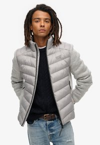 Gilet matelassé gris clair avec design matelassé et fermeture zippée, associé à un pull en tricot sombre et des manches grises, porté sur un jean bleu.
