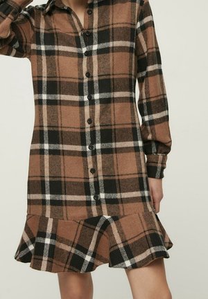 Bruine, zwarte en witte geruite blousejurk met lange mouwen en een uitlopende zoom, gedragen door een persoon tegen een effen lichte achtergrond.