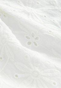 Tissu blanc avec des motifs brodés floraux et de petits détails découpés, montrant la texture et des coutures délicates.