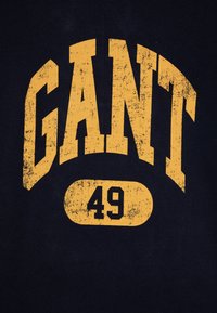 Námornícky modrý sveter s textúrovaným žltým potlačou s nápisom "GANT" a číslom "49" v oválnom ráme dole. Bavlnená látka, pohodlný dizajn.