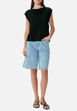 Femme portant un haut noir sans manches, un short en jean bleu clair jusqu’aux genoux et des sandales noires à brides, debout avec une main dans la poche.