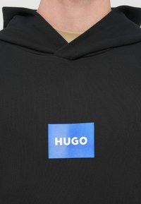 Čierna mikina s kapucňou s modrým obdĺžnikovým logom obsahujúcim biely nápis "HUGO" umiestneným na strede hrudníka.
