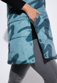Gestepptes Weste in Teal mit camouflage Muster, verfügt über ein schlankes Design, Druckknöpfe und Seitentaschen, aus einem glänzenden, leichten Material gefertigt.
