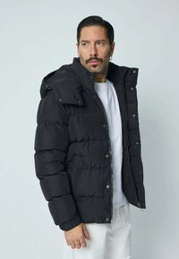 Doudoune noire à fermeture éclair sur le devant, avec capuche et poignets élastiques. Elle présente des panneaux verticaux cousus et des poches latérales pour le rangement.