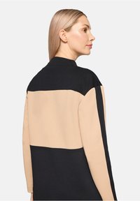 Kurtka w kolorze czarnym i beżowym z wysokim kołnierzem i długimi rękawami. Górna połowa jest czarna, a dolna beżowa, wykonana z miękkiego materiału.