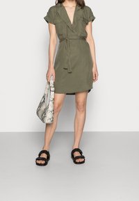 Abito avvolgente verde oliva realizzato in tessuto leggero, con maniche corte, una cintura e una scollatura a V profonda. Abbinato a sandali neri e una borsa decorata.