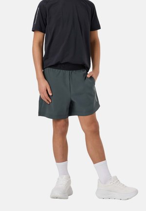 Personne portant un short de sport vert foncé, un t-shirt noir, des chaussettes blanches et des baskets blanches, debout avec une main dans la poche.