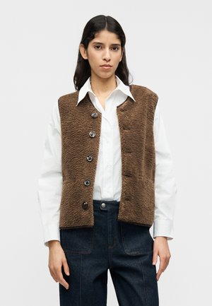 Καφέ υφασμάτινο γιλέκο με μπροστινό κούμπωμα με έξι μαύρα κουμπιά, φορεμένο πάνω από λευκό πουκάμισο με γιακά και συνδυασμένο με σκούρο μπλε παντελόνι.