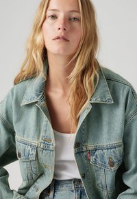 Lichtblauwe denimjack met een ontspannen pasvorm, zichtbare stiksels, twee voorzakken en een merkembleem op de borst. Gestructureerde stof.
