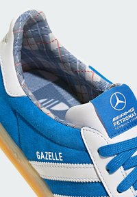 Baskets Adidas Gazelle bleu et blanc avec doublure intérieure à carreaux et logo de l’équipe AMG Petronas Formula 1 sur la languette.