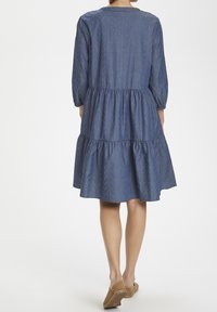 Žena v modré, vzorované, třístupňové šatech po kolena s 3/4 rukávy a béžovými nazouvacími botami, stojící na jednobarevném pozadí.