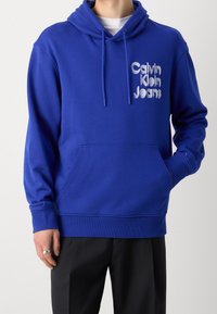 Blauwe katoenen hoodie met een voorzak in kangoeroestijl, een capuchon met trekkoord en een wit geborduurd "Calvin Klein Jeans" logo op de borst.