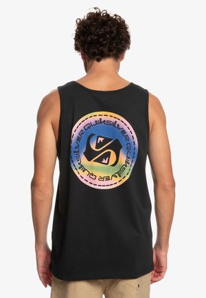 Homme aux cheveux bouclés portant un débardeur noir avec un logo circulaire coloré Quiksilver dans le dos, short beige, dos tourné.