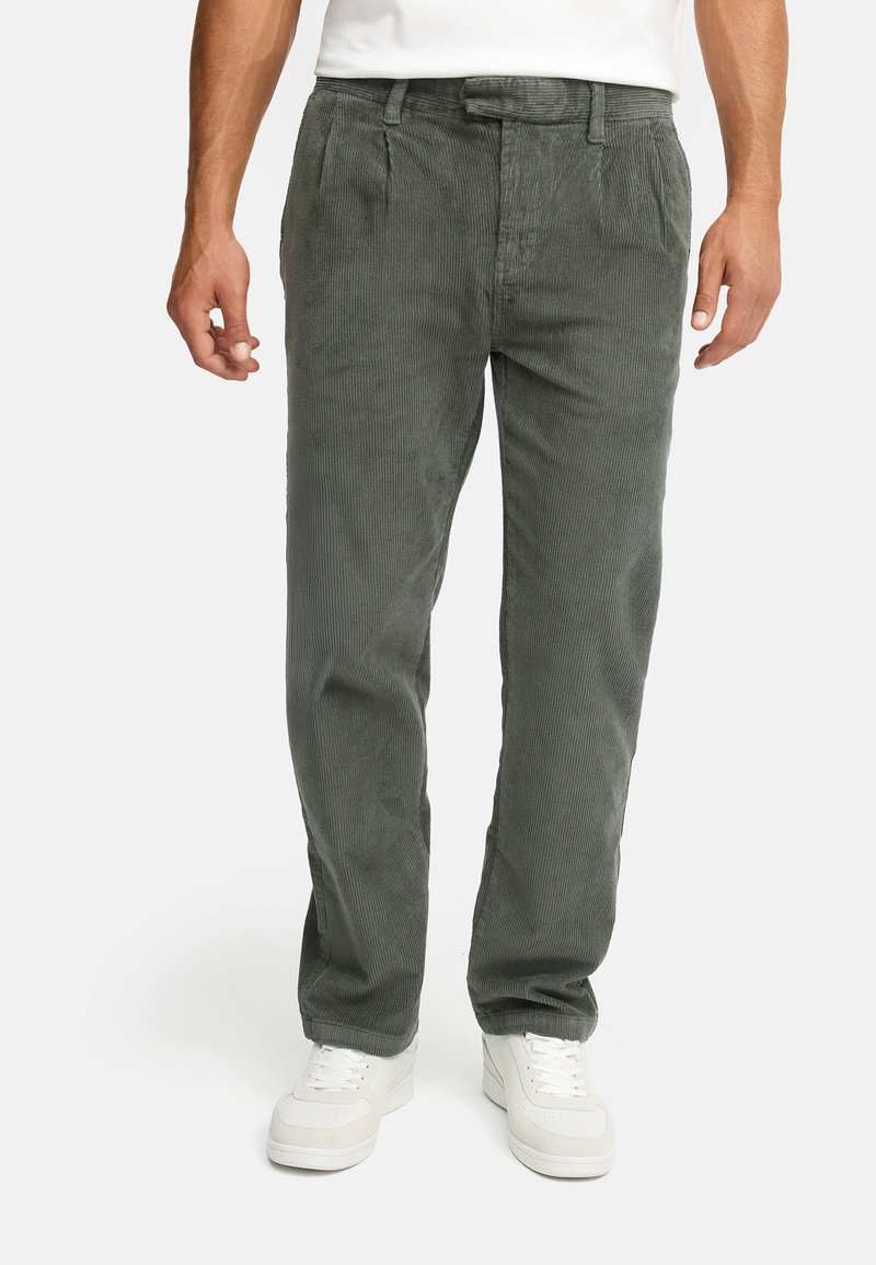 Homme portant un pantalon en velours côtelé gris avec des plis et des baskets blanches, debout devant un fond uni.