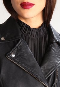 Giubbotto da moto in pelle nera con accessori in argento, caratterizzato da un design doppiopetto e un sottocollo a pieghe nera.