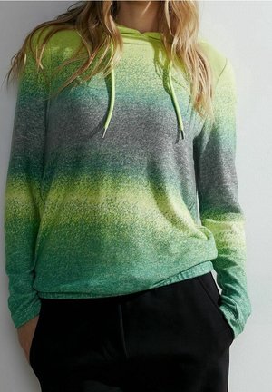 Femme portant un sweat à capuche dégradé vert, jaune et gris avec des cordons et un pantalon noir, mains dans les poches, debout devant un fond blanc.