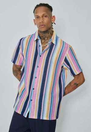 Homme avec des tatouages portant une chemise à manches courtes rayée verticalement colorée et un pantalon sombre, debout les mains dans le dos devant un fond uni.