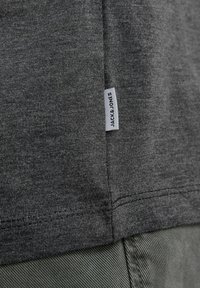 Camiseta gris con una tela suave y texturada. Presenta una pequeña etiqueta blanca con "JACK & JONES" cerca del dobladillo y costuras limpias a lo largo de los bordes.