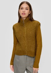 Strickweste in olivgrün mit hohem Kragen und halbem Reißverschluss. Begleitet von einem braunen Langarmshirt und karierten Hosen.